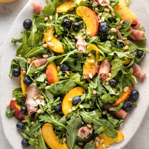 White platter with a peach arugula salad, with prosciutto, blueberries, pine nuts, and a white wine vinaigrette, ready to serve.
