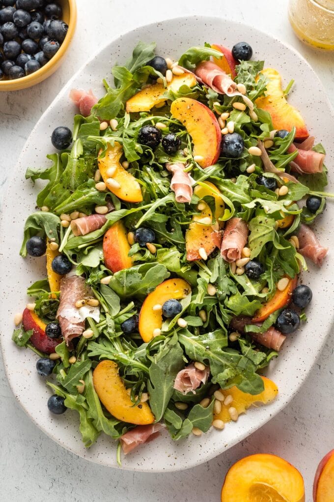 White platter with a peach arugula salad, with prosciutto, blueberries, pine nuts, and a white wine vinaigrette, ready to serve.