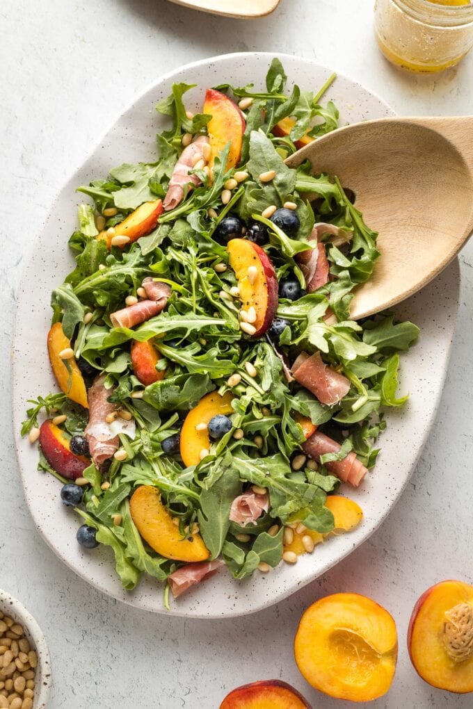 White platter with peach arugula salad and salad tongs preparing to serve portions.