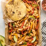 Sheet pan filled with fajita-seasoned chicken and veggies, surrounded by tortillas and small bowls of avocado and salsa.