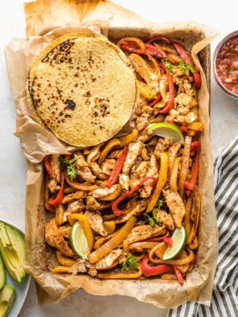 Sheet pan filled with fajita-seasoned chicken and veggies, surrounded by tortillas and small bowls of avocado and salsa.