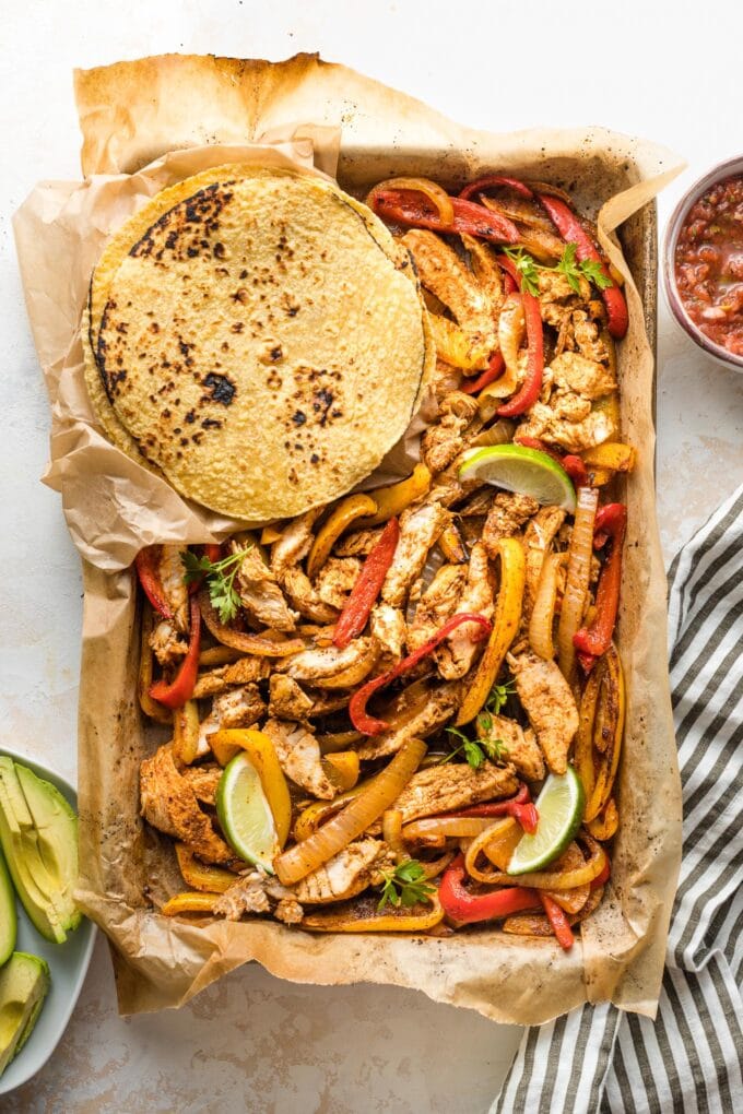 Sheet pan filled with fajita-seasoned chicken and veggies, surrounded by tortillas and small bowls of avocado and salsa.