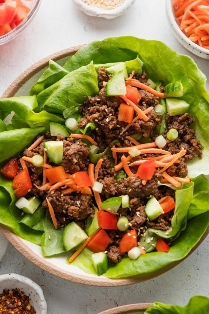 Small plate filled with three Korean beef lettuce wraps topped with fresh carrots, cucumber, bell pepper, green onions, and toasted sesame seeds.