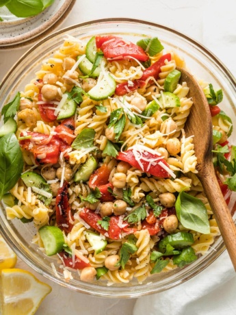 Clear serving bowl holding a pasta salad with chickpeas, roasted red peppers, cucumbers, and herbs.