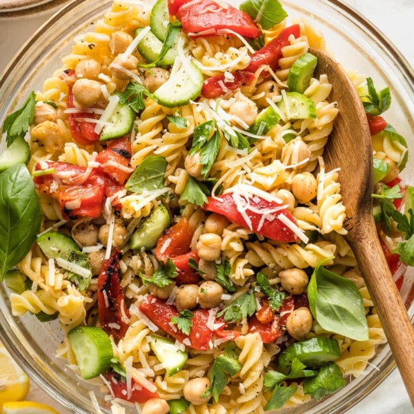 Clear serving bowl holding a pasta salad with chickpeas, roasted red peppers, cucumbers, and herbs.