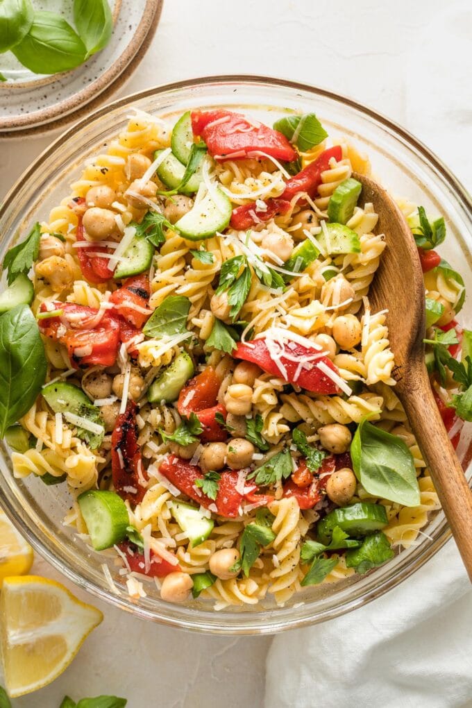Clear serving bowl holding a pasta salad with chickpeas, roasted red peppers, cucumbers, and herbs.