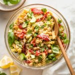 Clear serving bowl holding a pasta salad with chickpeas, roasted red peppers, cucumbers, and herbs.