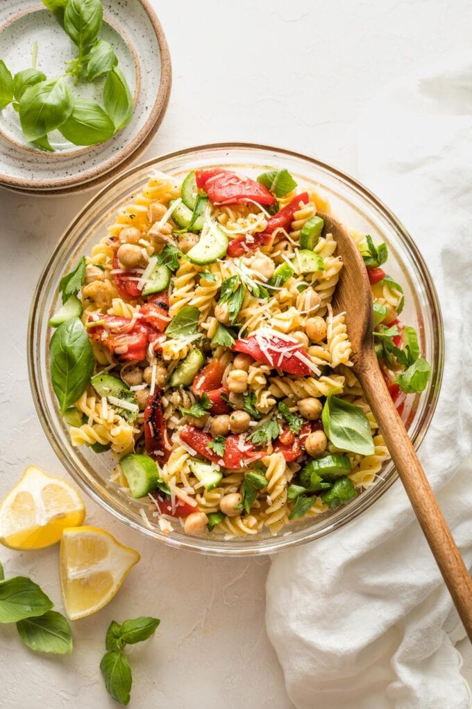 Clear serving bowl holding a pasta salad with chickpeas, roasted red peppers, cucumbers, and herbs.