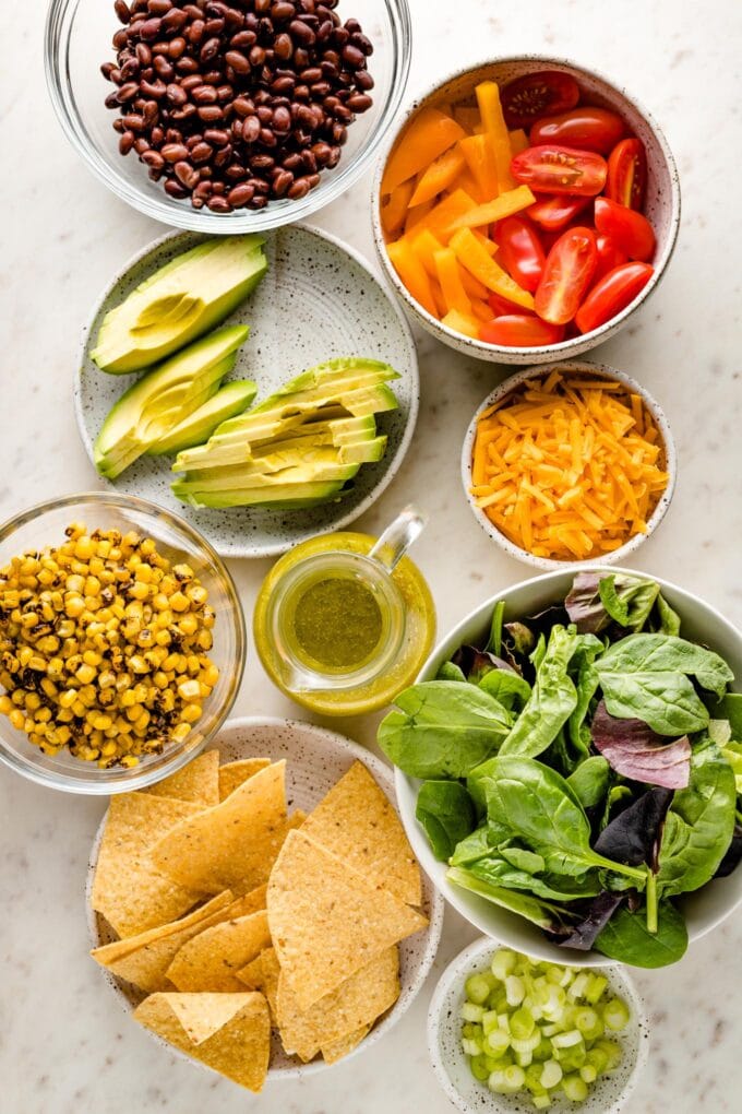 Prep bowls filled with ingredients: black beans, lettuce, bell pepper, cherry tomatoes, avocado, corn, cheese, and spices.