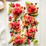 White platter with wedges of watermelon piled with fresh raspberries, mint, sea salt, lime juice, and balsamic glaze.