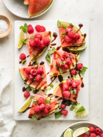 White platter with wedges of watermelon piled with fresh raspberries, mint, sea salt, lime juice, and balsamic glaze.