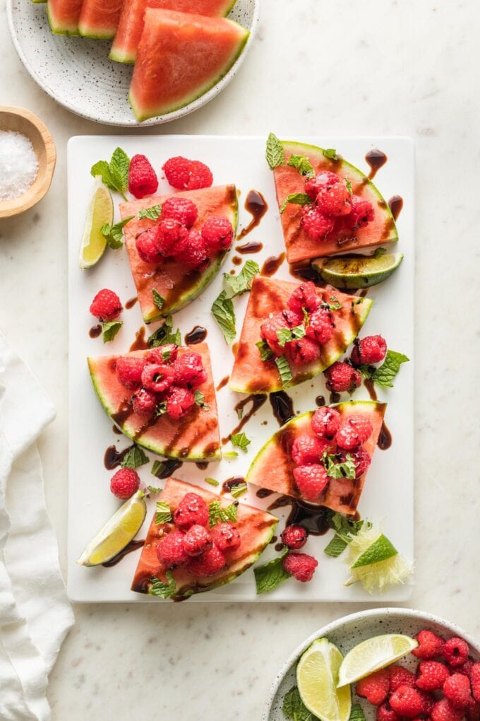 White platter with wedges of watermelon piled with fresh raspberries, mint, sea salt, lime juice, and balsamic glaze.