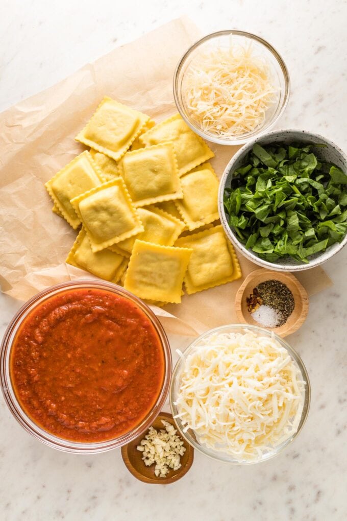 Uncooked ravioli, mozzarella and Parmesan cheese, fresh spinach, marinara sauce, garlic and spices arranged in prep bowls.