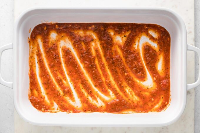 Marinara sauce spread in a thin layer in a large white rectangular baking pan.