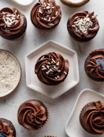 Large spread of chocolate cupcakes with a mix of chocolate frosting, white sprinkles, and blue sprinkles nearby.