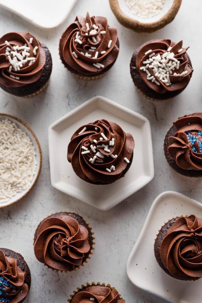 Large spread of chocolate cupcakes with a mix of chocolate frosting, white sprinkles, and blue sprinkles nearby.