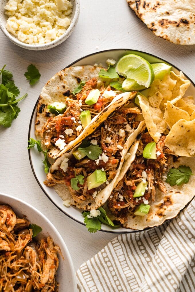 Small plate holding three shredded chicken tacos, surrounded by extra tortillas, cotija, and chicken