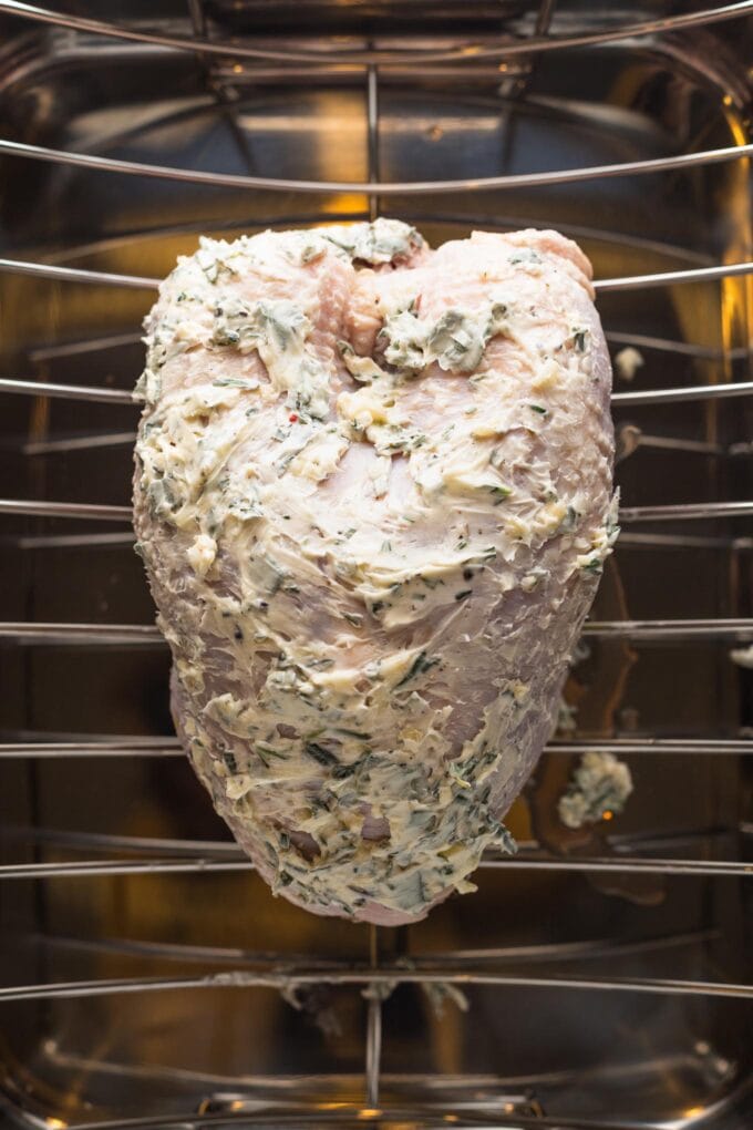 Turkey breast generously coated in a butter-garlic-herb mixture, set on a rack inside of a roasting pan.