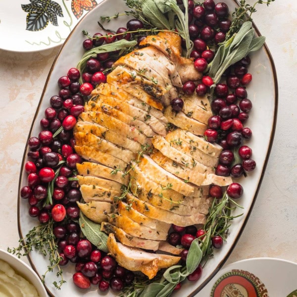Platter filled with an herb roasted turkey breast, sliced and ready to serve, garnished with cranberries and fresh herbs, served with gravy and mashed potatoes.