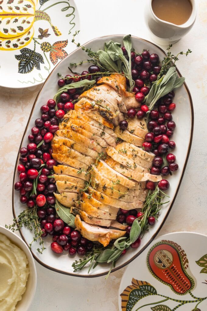 Platter filled with an herb roasted turkey breast, sliced and ready to serve, garnished with cranberries and fresh herbs, served with gravy and mashed potatoes.