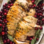Platter filled with an herb roasted turkey breast, sliced and ready to serve, garnished with cranberries and fresh herbs.