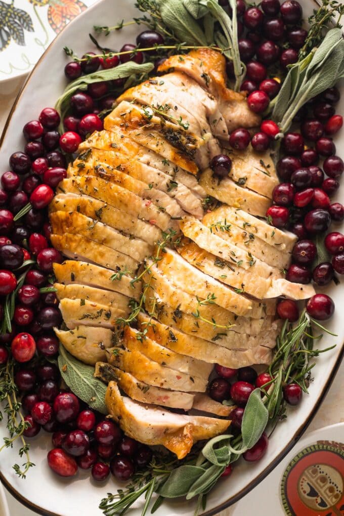 Platter filled with an herb roasted turkey breast, sliced and ready to serve, garnished with cranberries and fresh herbs.