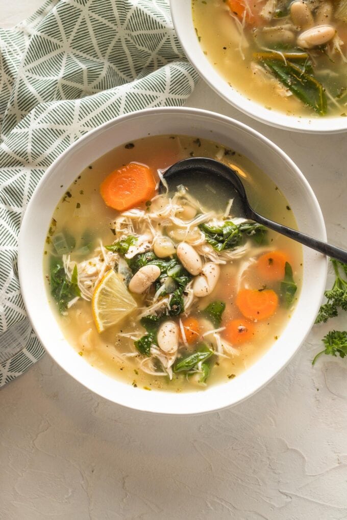 Close-up view of a bowl of white bean chicken soup with carrots and chard, with a spoon hovering above about to dig in.