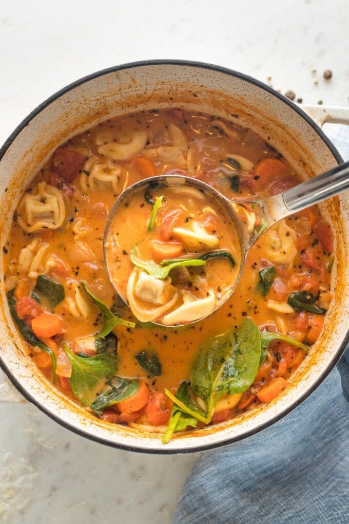 Finished creamy tomato tortellini soup being served with a silver ladle out of a white Dutch oven.