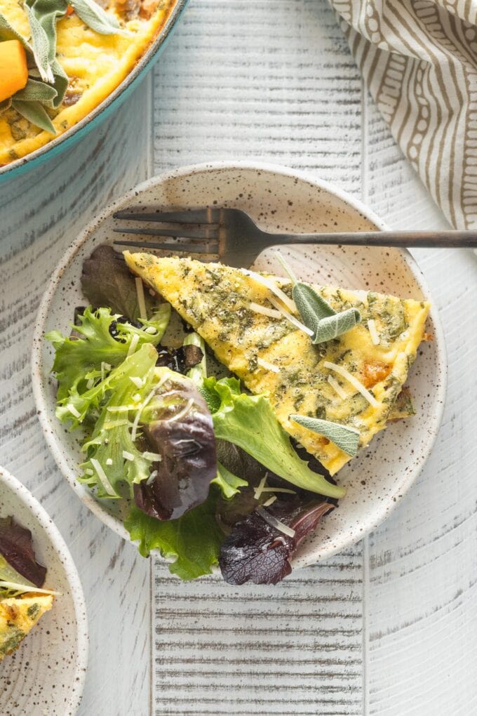 A generous slice of butternut squash frittata served on a small plate alongside baby greens.