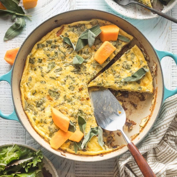 A butternut squash frittata with sage and red onion, sliced and being served from a cast iron skillet.