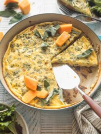 A butternut squash frittata with sage and red onion, sliced and being served from a cast iron skillet.