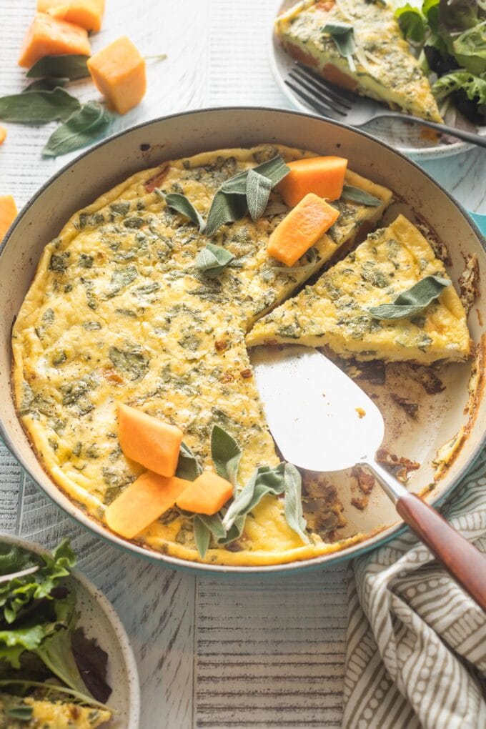 A butternut squash frittata with sage and red onion, sliced and being served from a cast iron skillet.