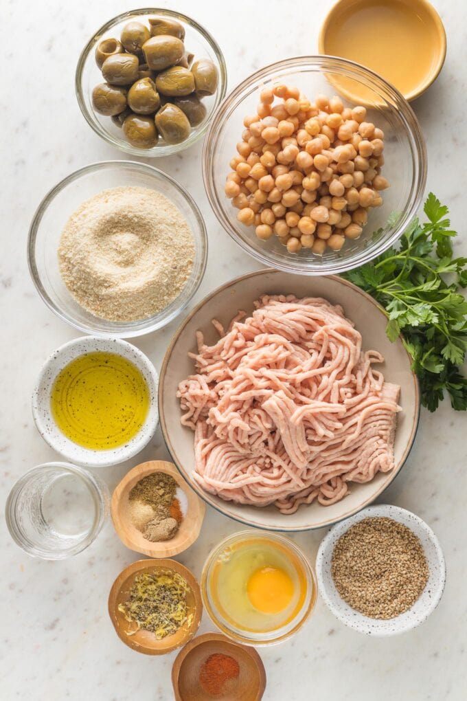 Prep bowls holding ground chicken, drained canned chickpeas, breadcrumbs, olives, sesame seeds, lemon juice, and seasonings.