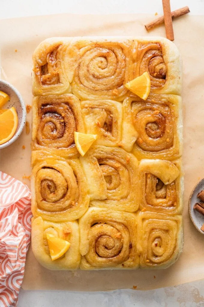 Glazed orange sweet rolls just tilted out of a baking pan onto parchment and garnished with extra orange zest and wedges.