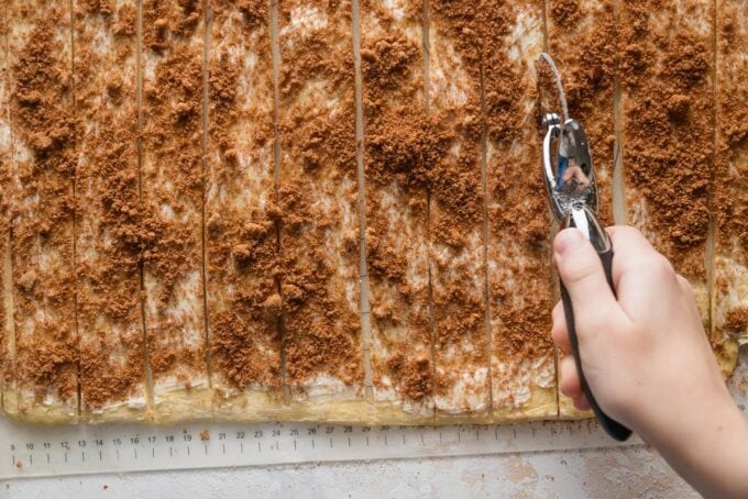 A child's hand using a pizza wheel to cut a sheet of cinnamon roll dough into individual strips.