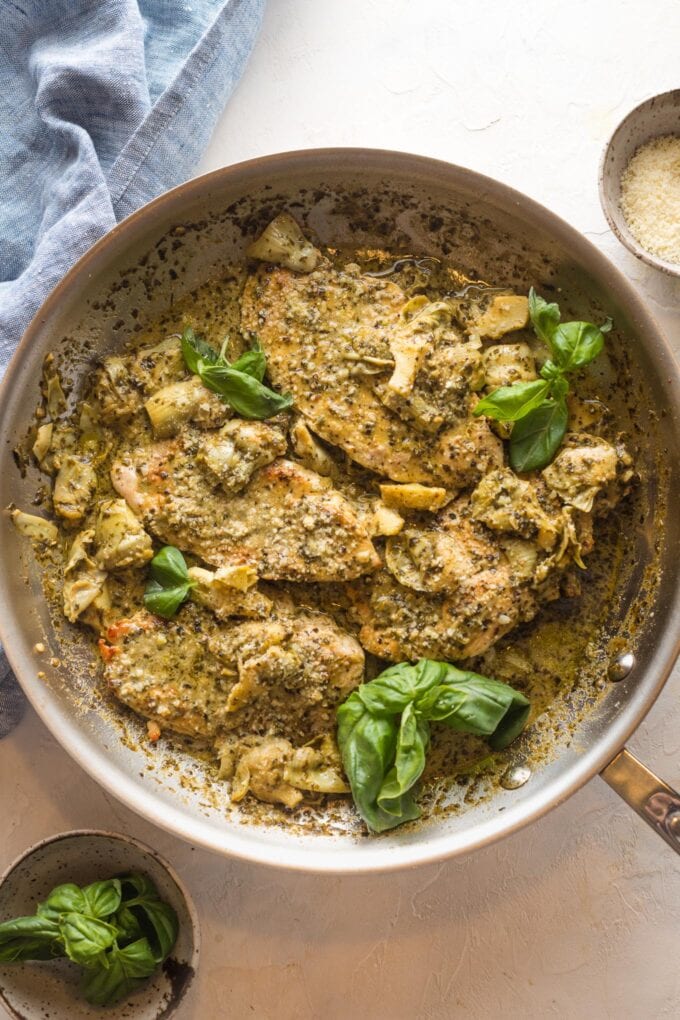 Skillet filled with creamy pesto artichoke chicken served with Parmesan and fresh basil leaves.