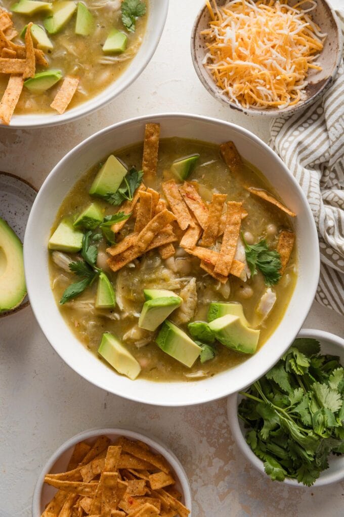 Overhead view of a bowl full of white bean salsa verde chicken chili, with extra toppings nearby on the counter.