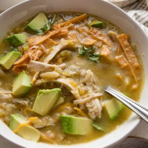 Close up of a bowl of white bean chicken chili with salsa verde topped with avocado, tortilla strips, and shredded cheese.