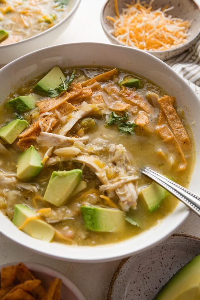 Close up of a bowl of white bean chicken chili with salsa verde topped with avocado, tortilla strips, and shredded cheese.