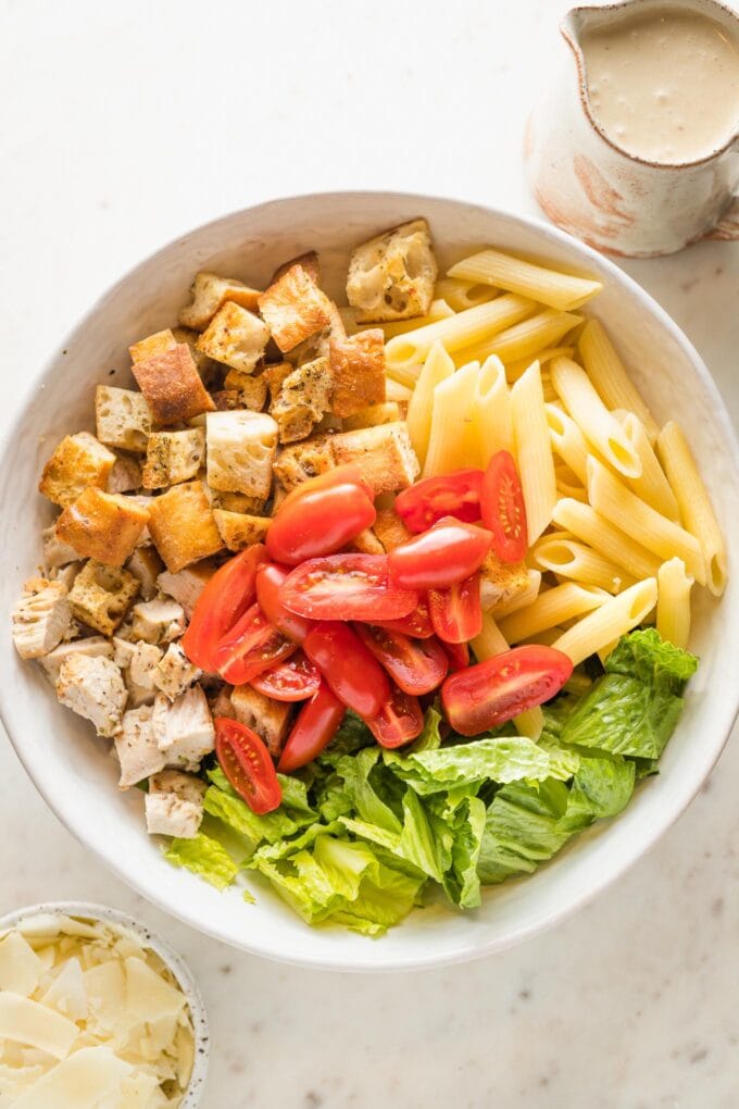 Romaine, penne, tomatoes, croutons, and chicken breasts in a large bowl.