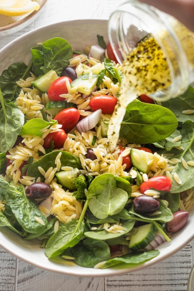 Dressing being poured out of a jar and onto a salad with orzo and spinach.