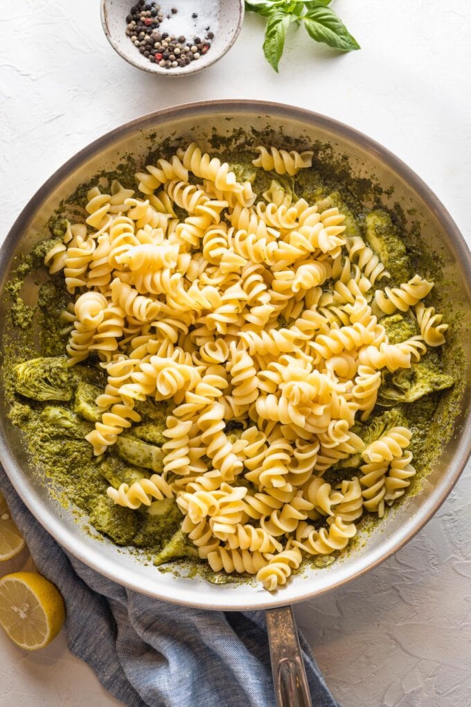 Pasta being added to a skillet with pesto cream sauce and artichoke hearts.