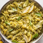 Large skillet full of pasta with artichoke hearts, pesto, and lemon wedges.