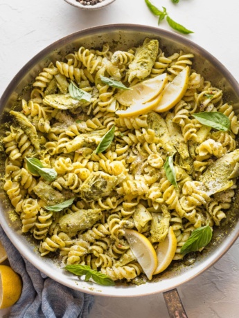 Large skillet full of pasta with artichoke hearts, pesto, and lemon wedges.