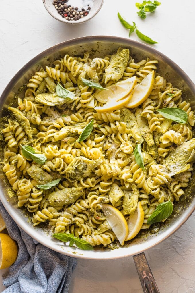 Large skillet full of pasta with artichoke hearts, pesto, and lemon wedges.
