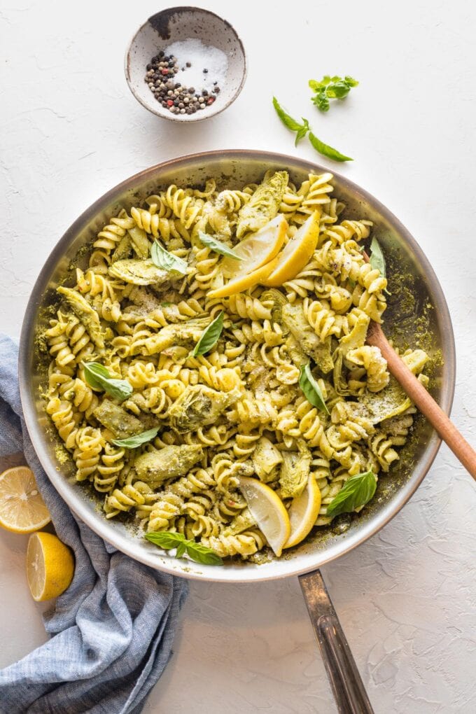 A serving spoon about to portion out helpings of pesto pasta with artichoke hearts.