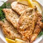 Close up of pan-fried Greek-marinated chicken breasts piled on a plate with fresh dill and lemon wedges.