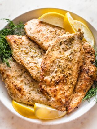 Close up of pan-fried Greek-marinated chicken breasts piled on a plate with fresh dill and lemon wedges.