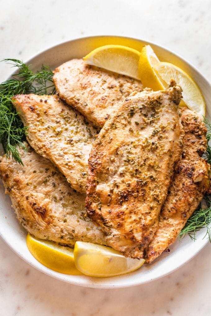 Close up of pan-fried Greek-marinated chicken breasts piled on a plate with fresh dill and lemon wedges.