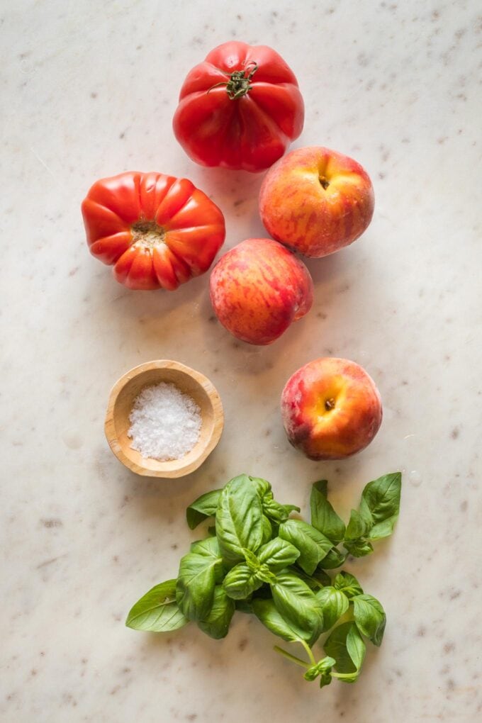 Heirloom tomatoes, peaches, sea salt, and fresh basil leaves scattered on a white countertop.